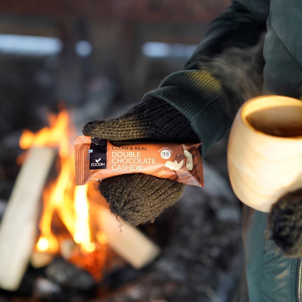 Wearing dark gloves, a person holds a Foodin Clean & Real Protein Bar Double Chocolate Cashew and a wooden cup by a campfire with burning logs in the background.
