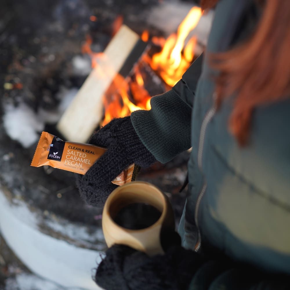 Someone in dark gloves holds a Foodin Clean & Real Protein Bar Salted Caramel Pecan and a wooden cup near an outdoor fire pit with burning logs.