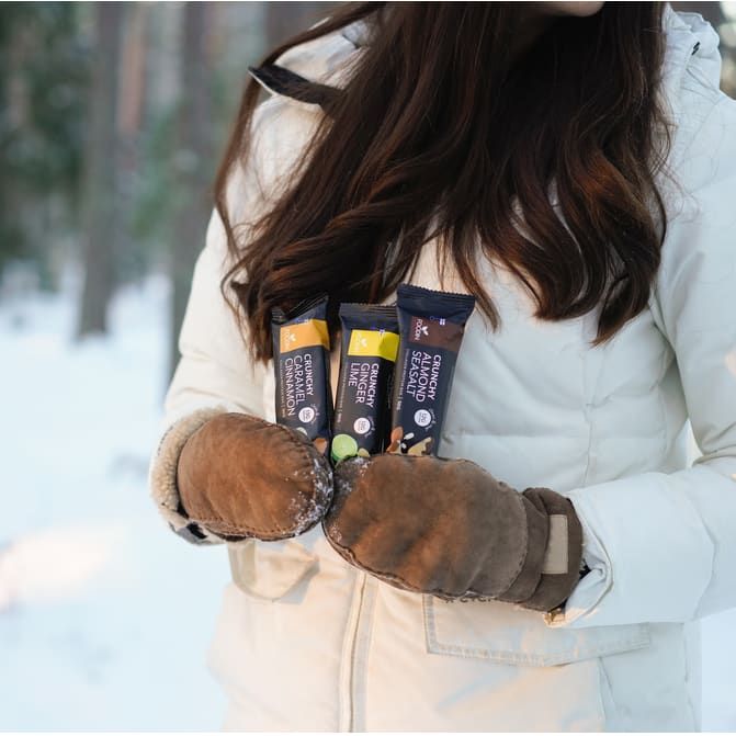 A woman in a white winter coat and brown mittens stands in a snowy forest, holding three Foodin Collagen Protein Bars, including the Crunchy Caramel Cinnamon flavor.