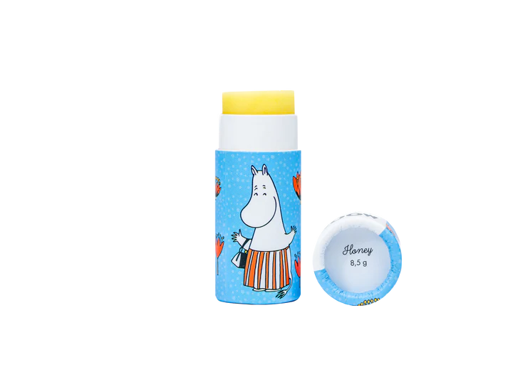 A tube of Gustaf & Linnea Moomin Lipbalm Moominmamma with a blue wrapper, orange hearts, and a cartoon Moomin character. The cap is off, revealing the Honey 8.5g label inside. Eco-friendly product by Gustaf & Linnea.