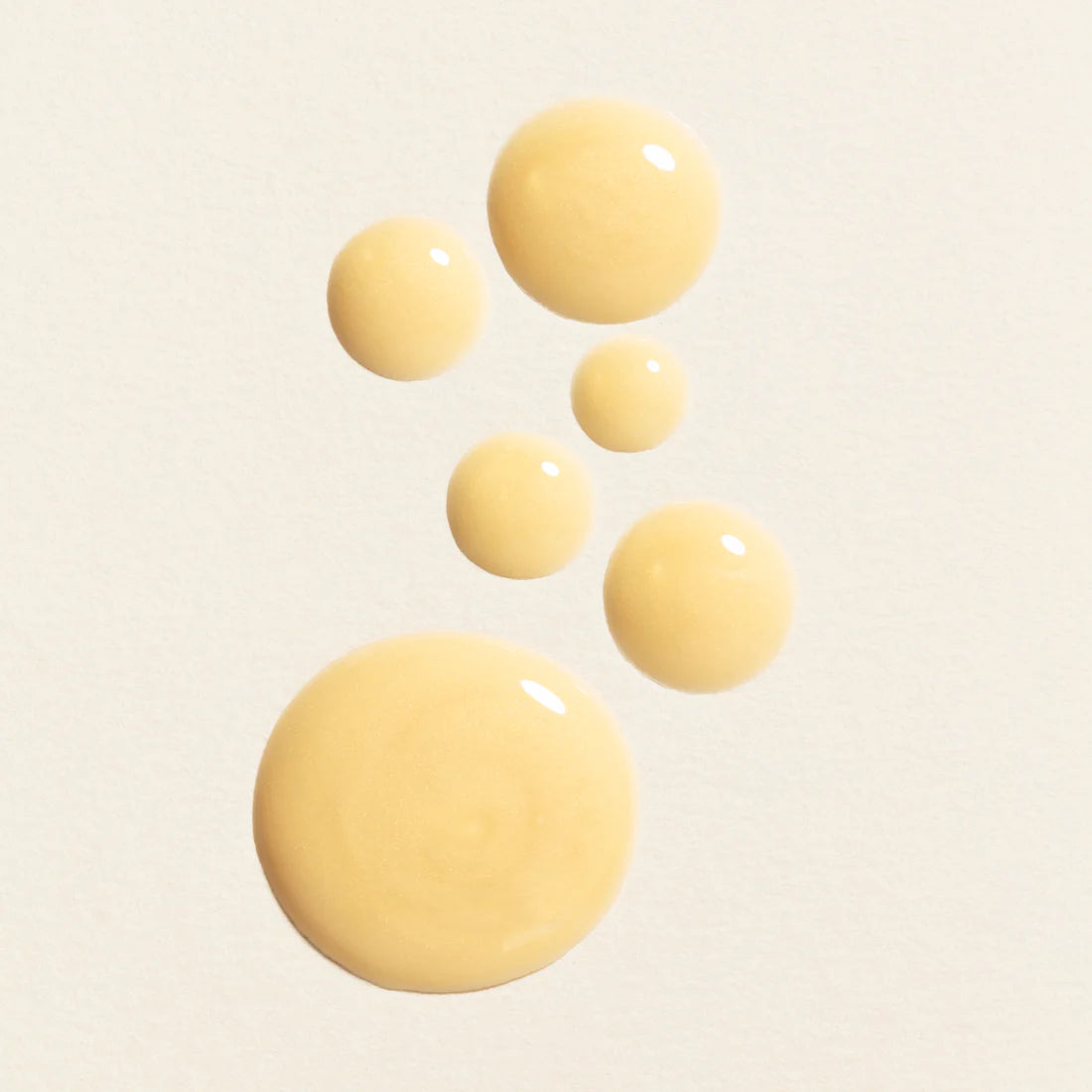 Six drops of Lumene Nordic-C Glow Boost Eye Serum are arranged in a loose, upward curve on a light, textured background. The drops differ in size, with the largest at the bottom and the smallest near the top.