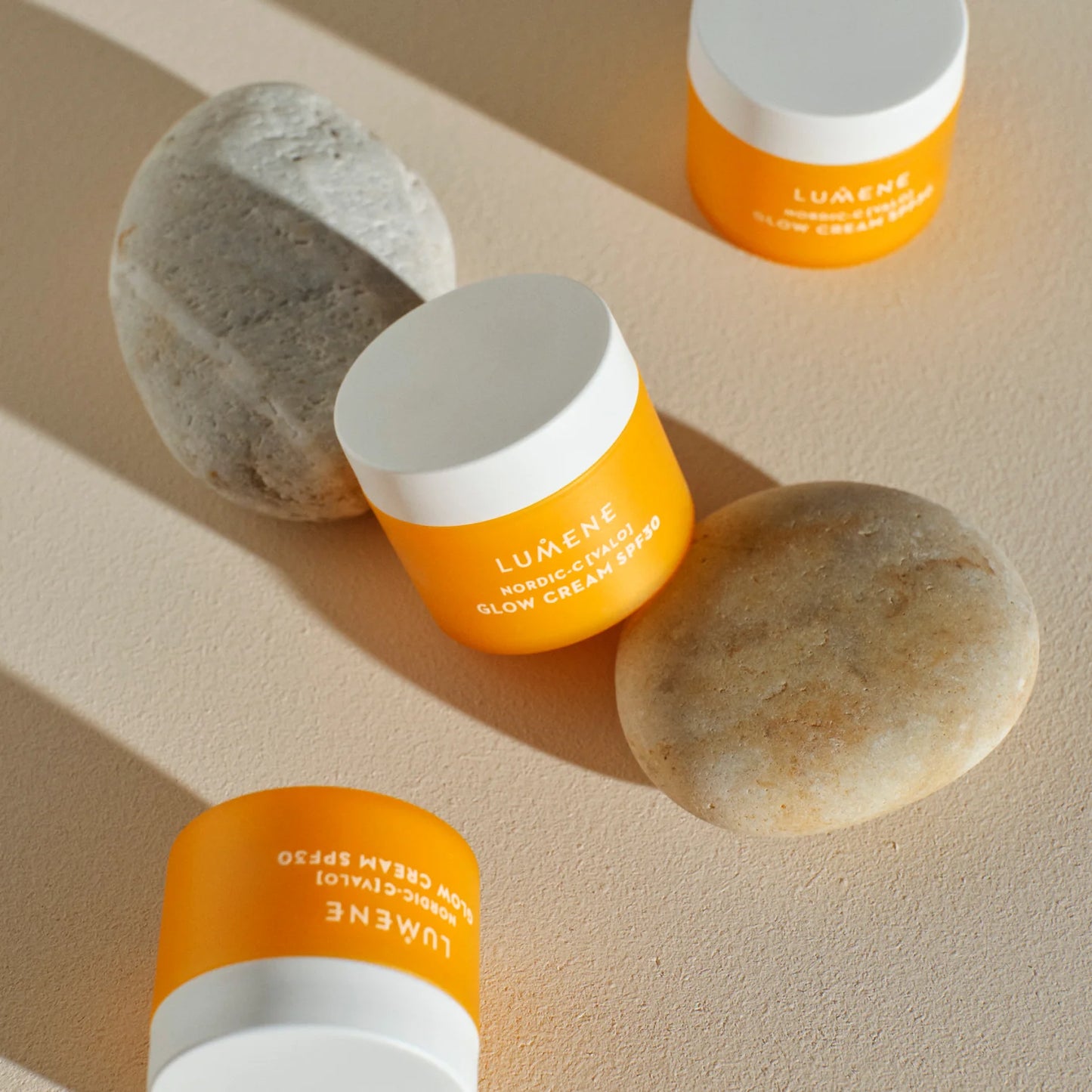 Four orange and white jars of Lumene Nordic-C Glow Cream SPF30 are arranged on a beige surface beside two smooth, light-colored stones, creating a minimalist and natural composition.