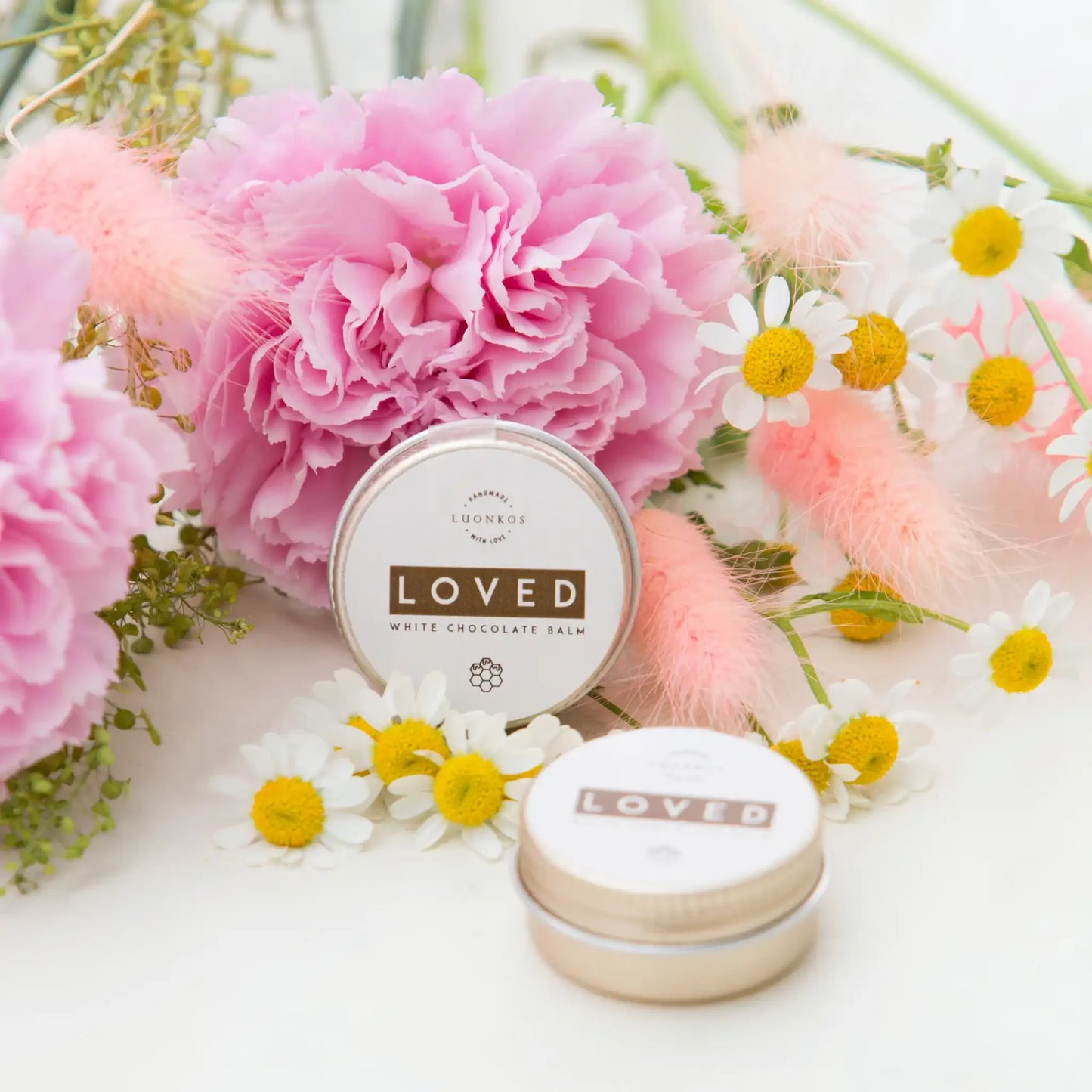 A tin of Luonkos Loved White Chocolate Multipurpose Lip Balm sits among pink carnations, daisies, and soft pink foliage, with another tin in front. The scene is bright, fresh, and floral.