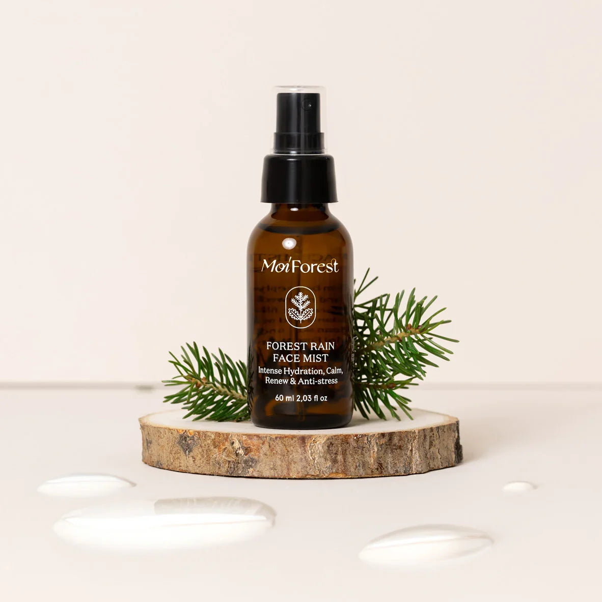 A bottle of Mayforest Forest Rain Face Mist in an amber glass bottle with a black spray top, styled on a wooden slice coaster with green pine branches, on a clean off-white background with decorative liquid droplets.