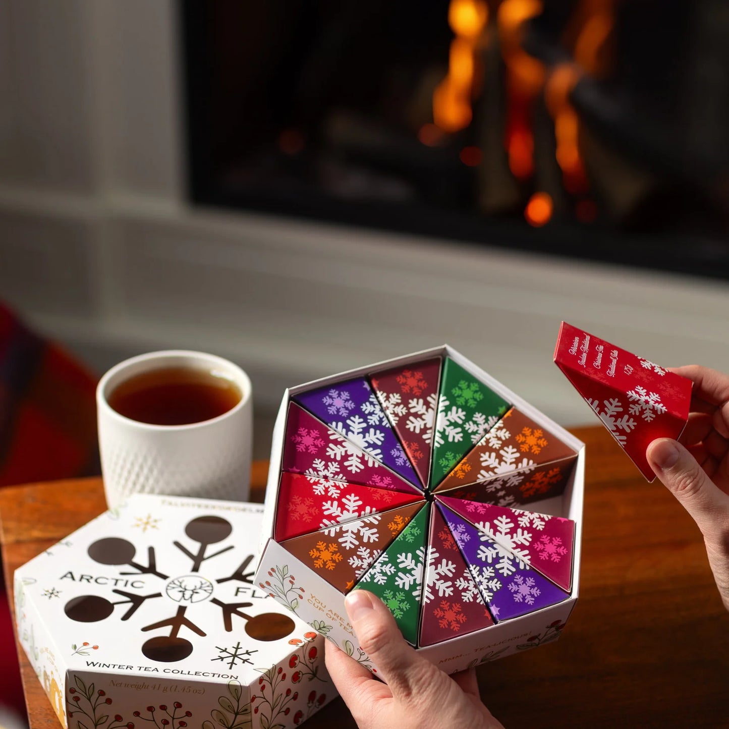 Someone holds a red packet from the Nordqvist Winter Tea Collection by Nordqvist, taken from a hexagonal gift box with colorful snowflakes. A cup of tea and fireplace in the background set a cozy holiday mood.