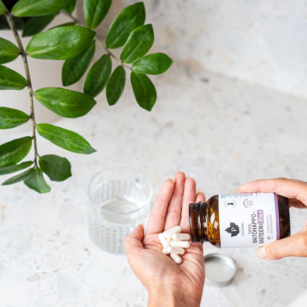 A hand holds white capsules of Puhdistamo Strong Probiotic 30 Billion CFU, poured from a Puhdistamo bottle over a marble countertop beside a glass of water and a leafy green plant.