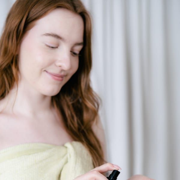 A woman with long brown hair, wrapped in a light yellow towel, smiles gently as she dispenses Rimita Green's RimitaGold 100% Virgin Argan Oil from a pump bottle onto her hand. White curtains hang in the background.