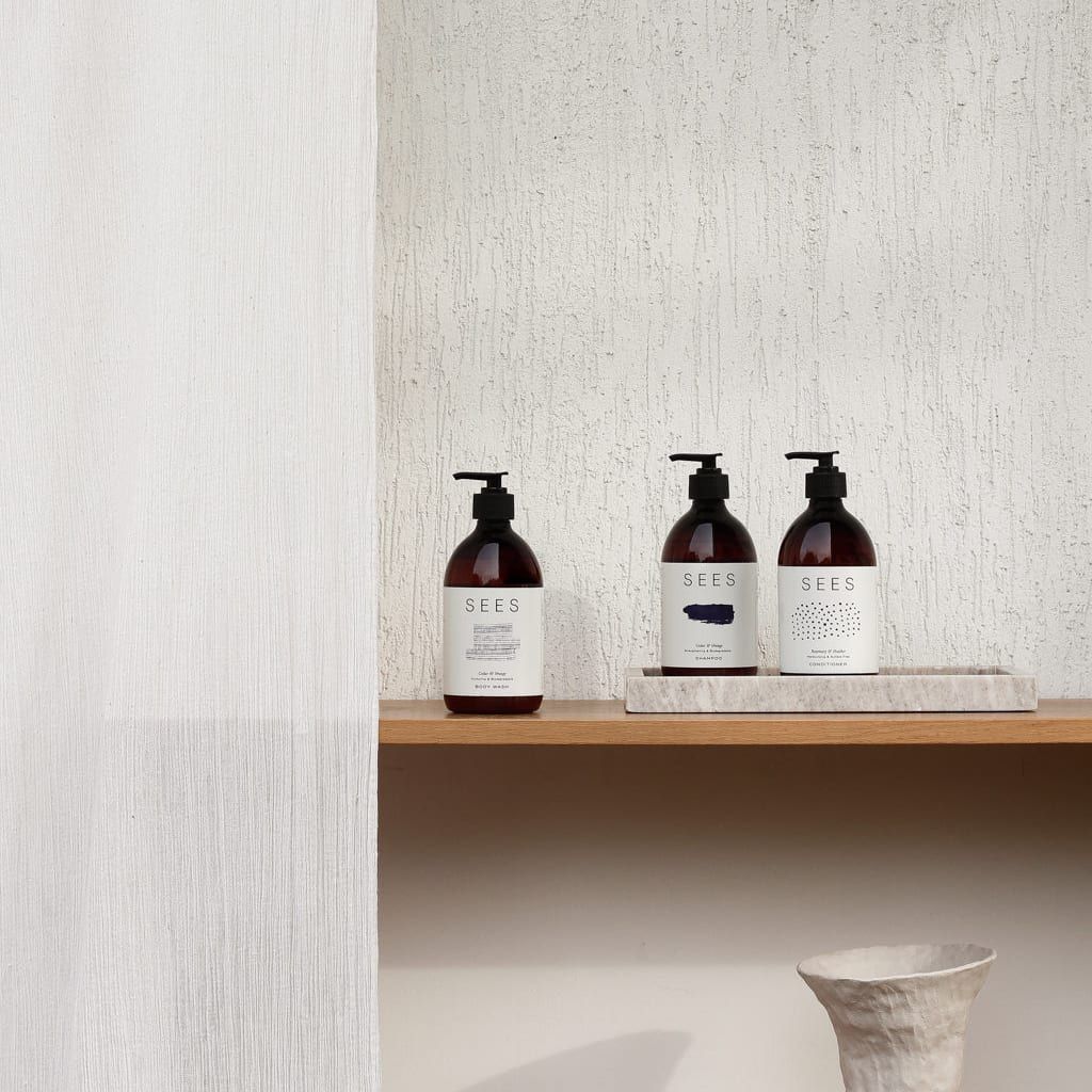 Three amber pump bottles of SEES Company’s SEES Body Wash, Cedar & Orange, with minimalist labels, are arranged on a wooden shelf against a textured white wall beside a white curtain and a pale ceramic vase below the shelf.