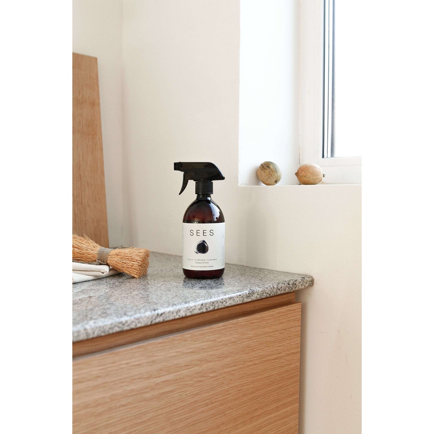 A SEES Company Cleaning Spray for the Kitchen and Bathroom sits on a kitchen countertop near a window, beside a cleaning brush and decorative stones—ideal for anyone seeking a plant-based cleaning spray.
