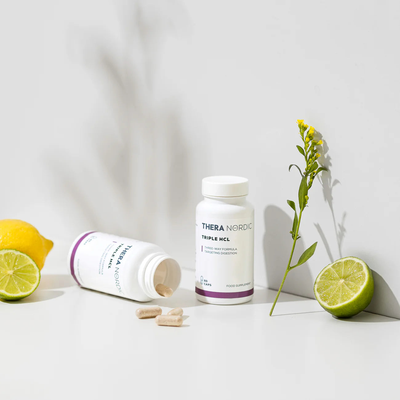 Two THERA Nordic Triple HCL bottles from THERA Nordicâone upright and one tipped with tablets spilling outâare shown with a lemon, halved lime, and small yellow flower, highlighting this digestive health supplement on a light background.