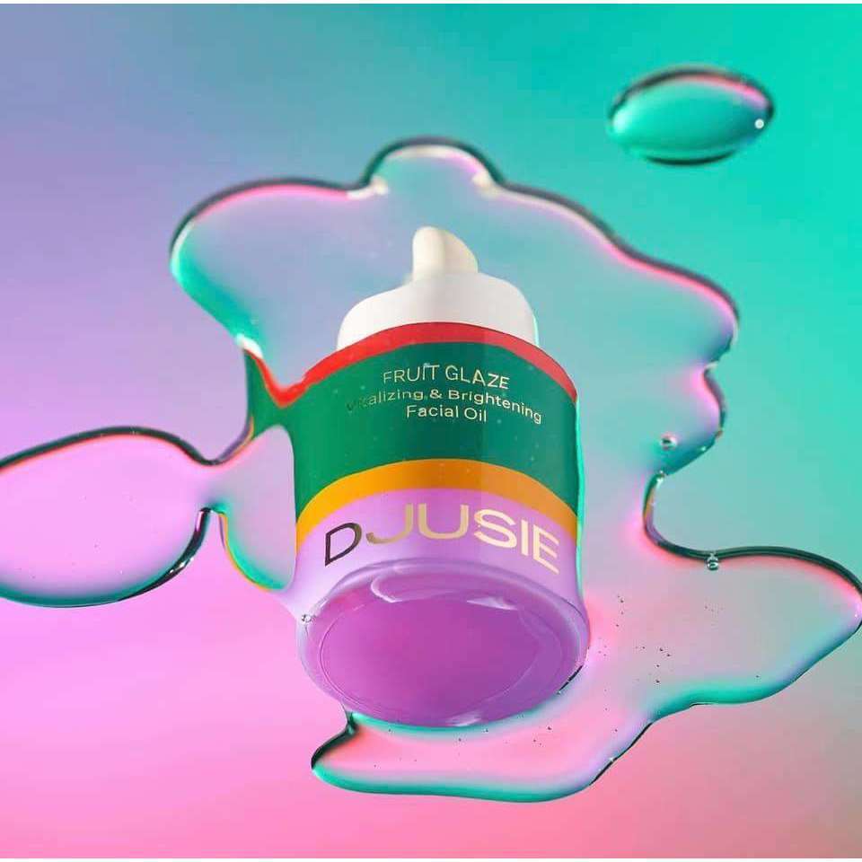 A dropper bottle of Djusie Fruit Glaze Vitalizing & Brightening Face Oil sits on a colorful, iridescent surface with oil puddles reflecting pink, green, and blue—ideal for glowing skin and pigment spot prevention.