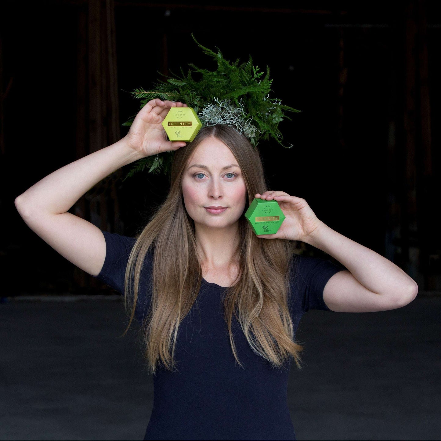 A woman with long blonde hair wears a crown of greenery and holds a yellow hexagon and the Luonkos Infinity Oil Cleansing Cake, ideal for sensitive skin, near her head against a dark background.