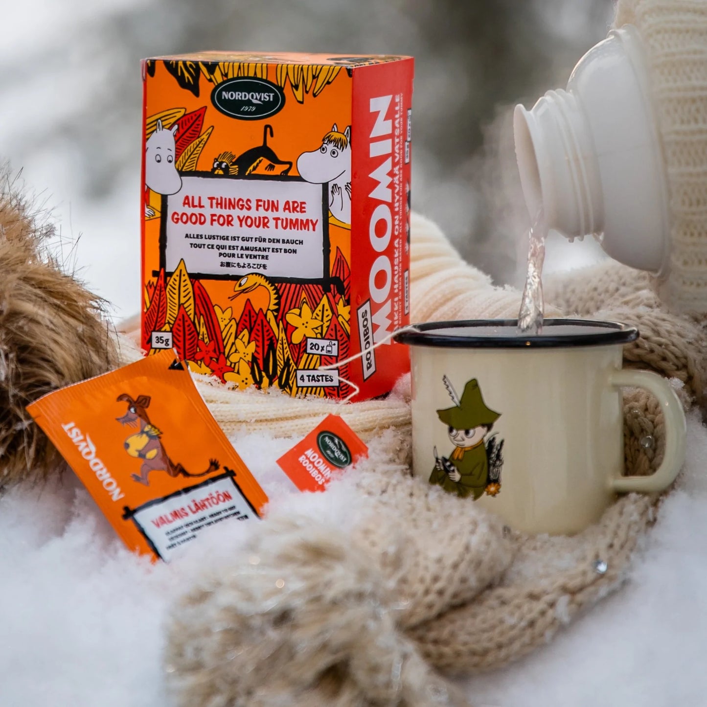 Wrapped in warm winter clothes, someone pours a hot drink from a thermos into a Snufkin mug beside a box and packets of Nordqvist All Things Fun Are Good For Your Tummy rooibos tea, all set on snow.