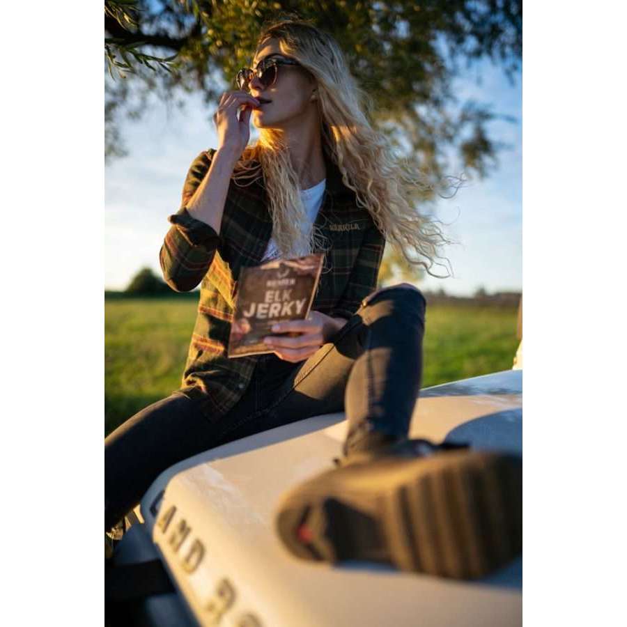A woman sits on a car hood enjoying Renjer Nordic Elk Jerky Black Pepper, savoring its smoky taste and hint of black pepper.