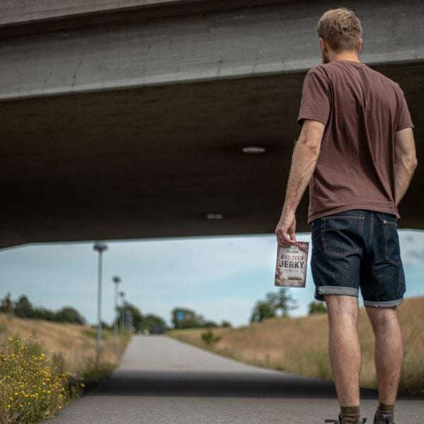 A man in a brown t-shirt and denim shorts walks on a path under a bridge, holding a bag of Renjer Nordic Red Deer Jerky Chili & Lime, with grassy fields and a streetlamp in the background.