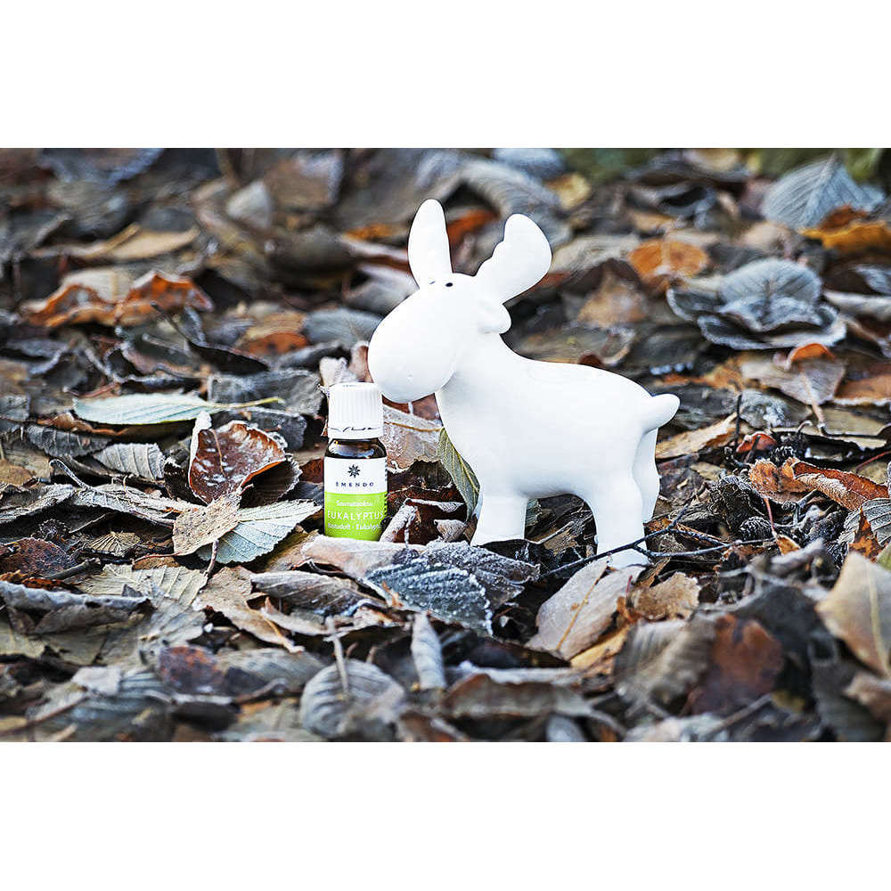 The Emendo Sauna Reindeer, a small white ceramic ornament, and Emendo Eucalyptus Oil rest on dry leaves, creating a charming sauna scene.