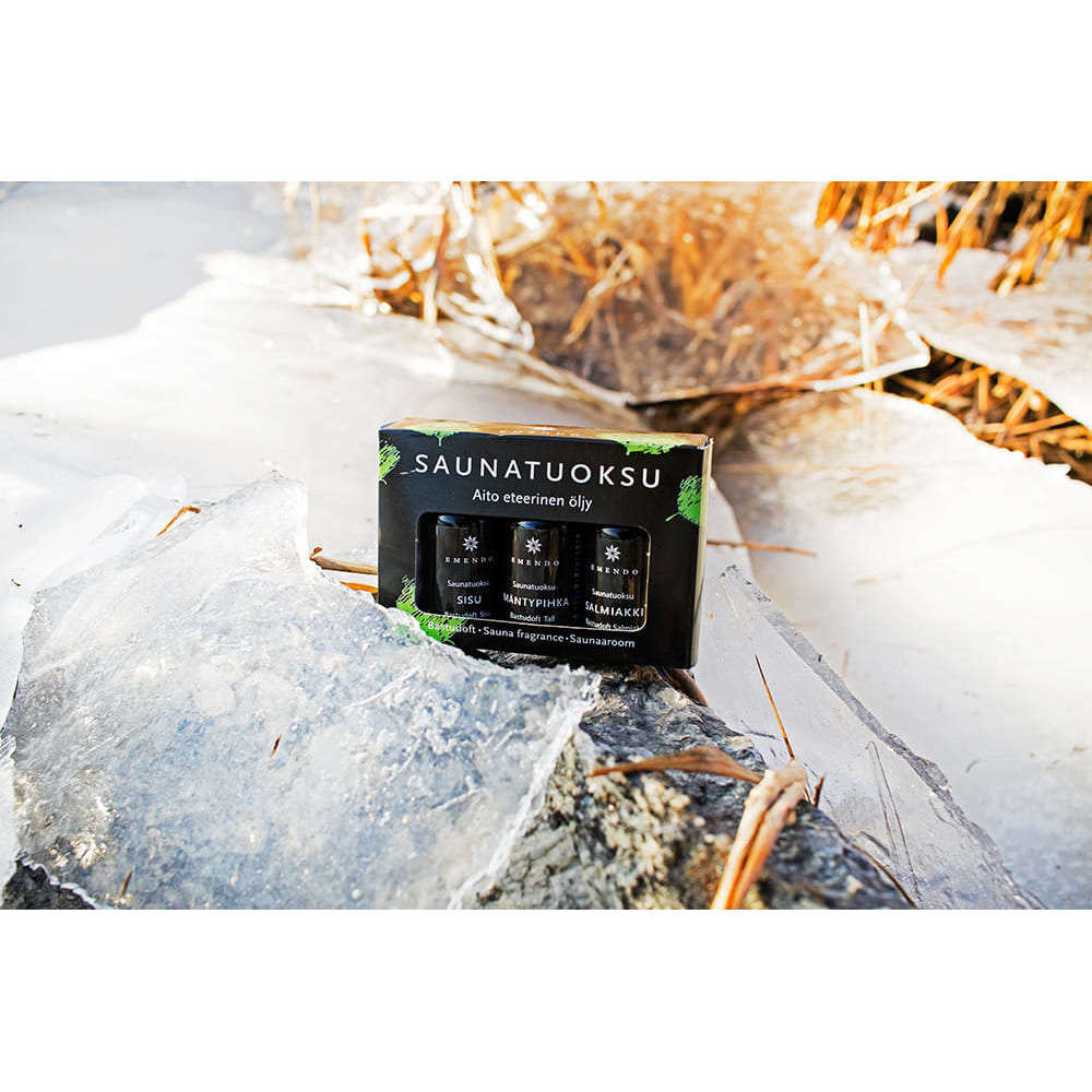 An Emendo Sauna Fragrance Salmiac, Pine & Sisu box by Emendo, made in Finland, rests outdoors on cracked ice among dried reeds with sunlight shining behind.