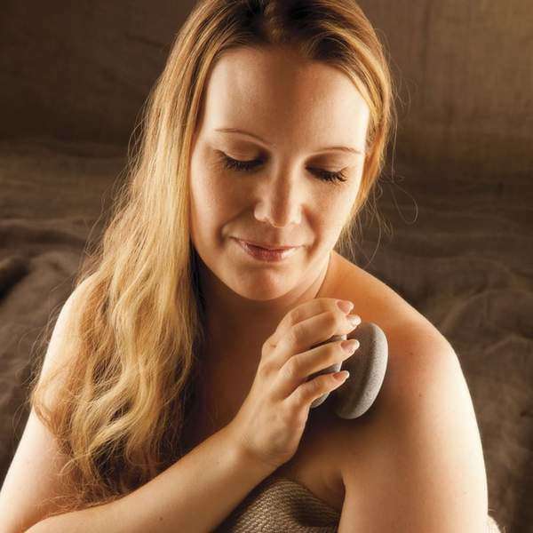 A woman with long blonde hair gently exfoliates her shoulder using the Hukka Sauna-Joy Massage Aid by Hukka, smiling softly and wrapped in a towel against a cozy, neutral background.