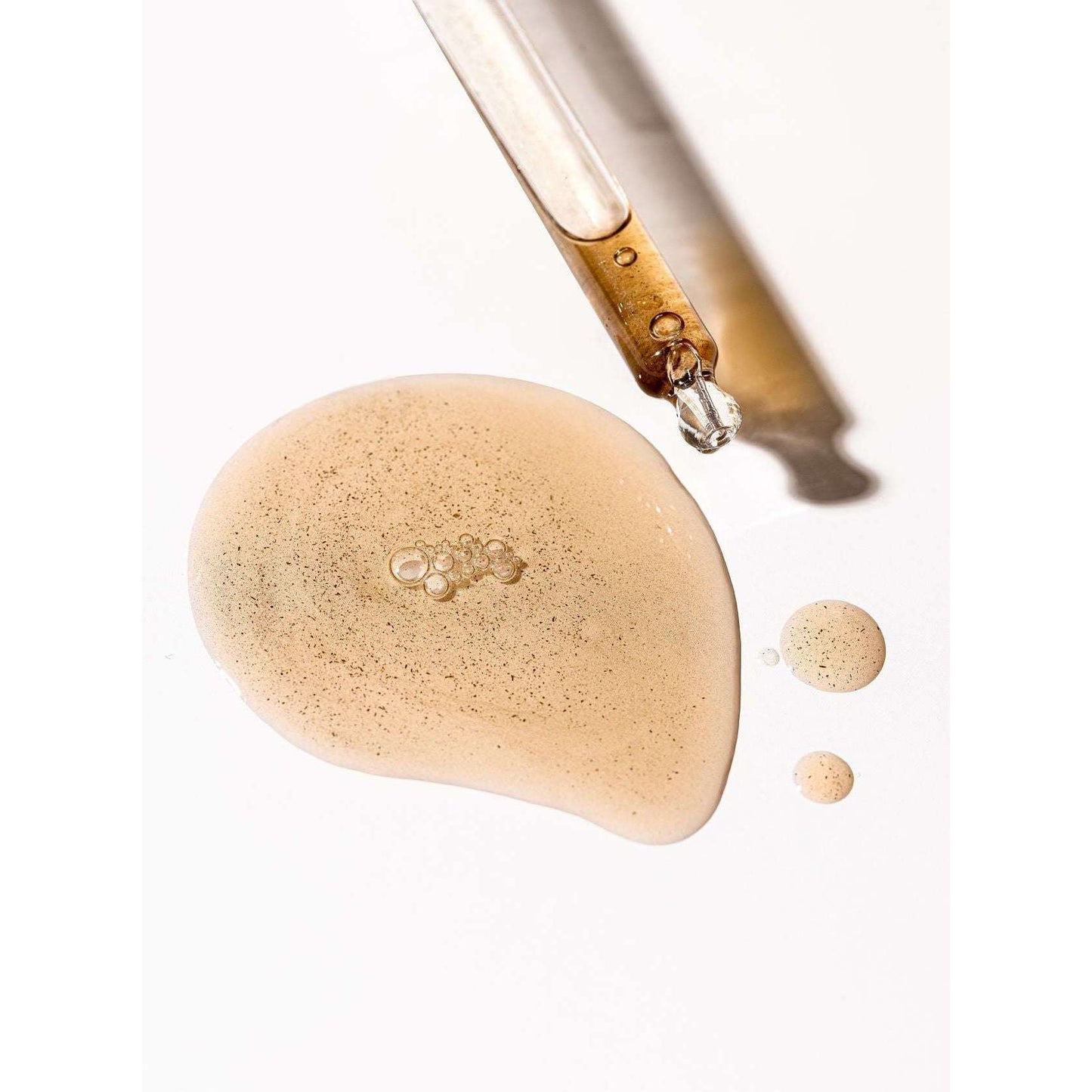 A dropper releases Moi Forest Dust Microbiome Magic Oil onto a white surface, creating one large and two small golden, shimmering droplets of this multipurpose skin oil by Moi Forest, each speckled with tiny particles.