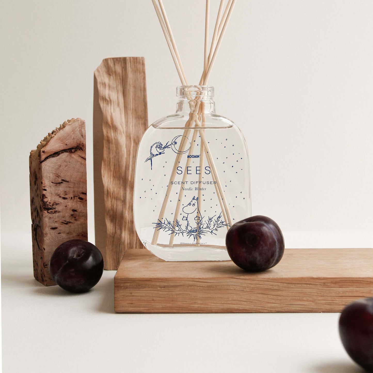 The SEES x Moomin Room Diffuser Nordic Winter by SEES Company rests on a light wooden surface with wooden blocks and plums, featuring delicate blue illustrations and capturing the essence of Finnish design in a neutral setting.