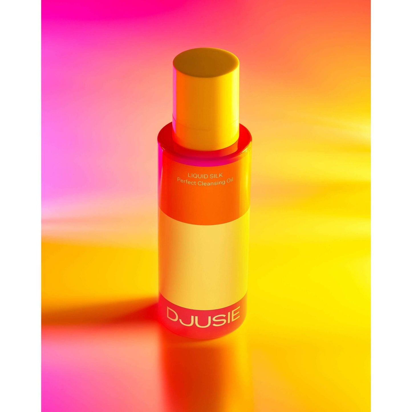A bottle of Djusie Liquid Silk Perfect Cleansing Oil stands upright against a vibrant yellow, orange, and pink gradient. The sleek bottle features a yellow cap and bold white branding, effortlessly removing water-resistant makeup.