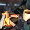 Wearing dark gloves, a person holds a Foodin Clean & Real Protein Bar Double Chocolate Cashew and a wooden cup by a campfire with burning logs in the background.
