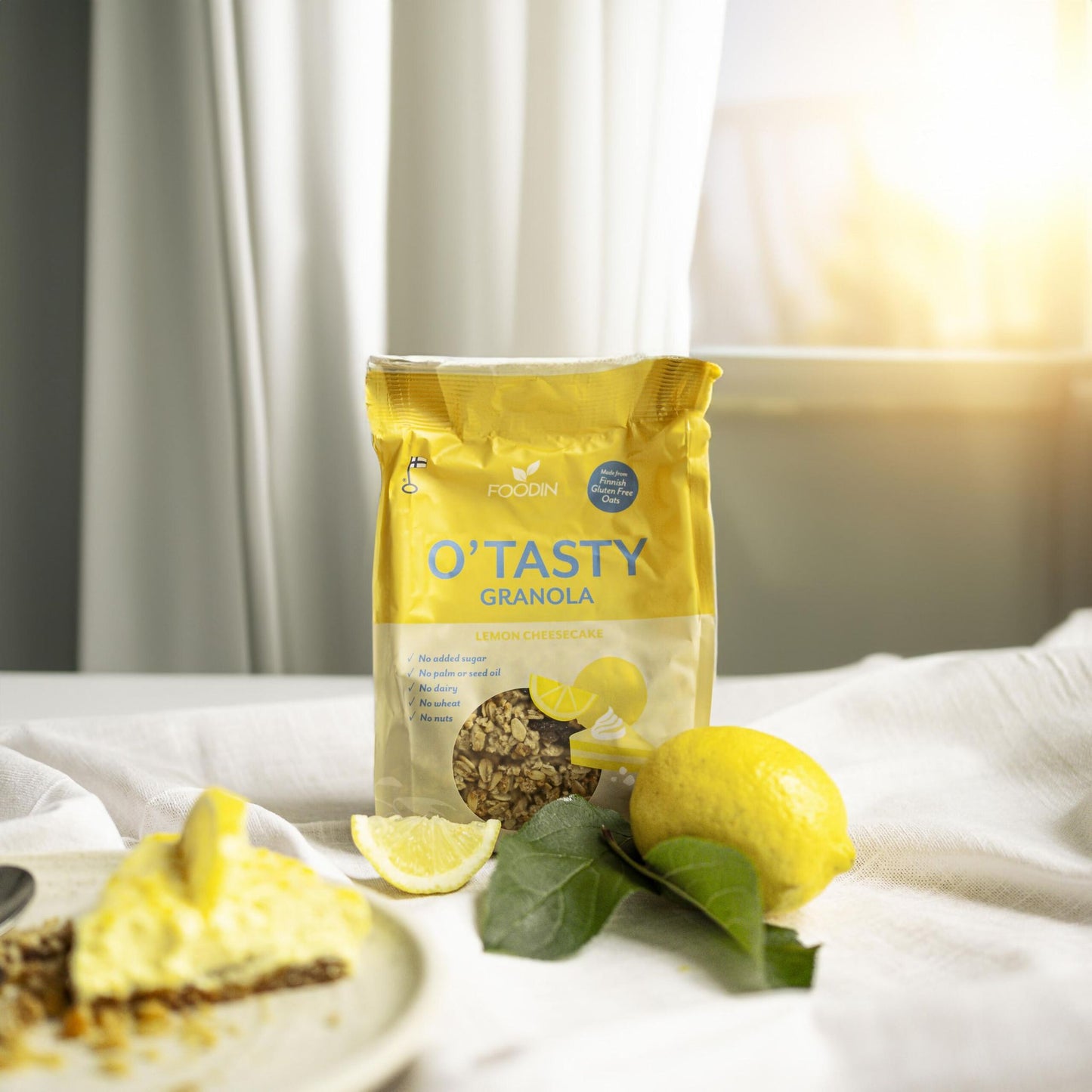 A bag of Foodin O`Tasty Kauragranola Lemon Cheesecake sits on a white tablecloth near a lemon, lemon wedge, green leaves, and cheesecake, all illuminated by sunlight through the window.
