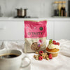 A bag of Foodin O`Tasty Kauragranola Raspberry Muffin sits on a white surface beside a coffee, a raspberry muffin with cream and raspberries, and scattered granola pieces. A modern kitchen appears blurred in the background.
