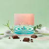 A square package of Caine & Balanced chocolate stands upright on a pale mint green surface with scattered dark chocolate pieces around it. The package has a soft peach-to-blue gradient with concentric ripple graphics and a small blue droplet in the center.