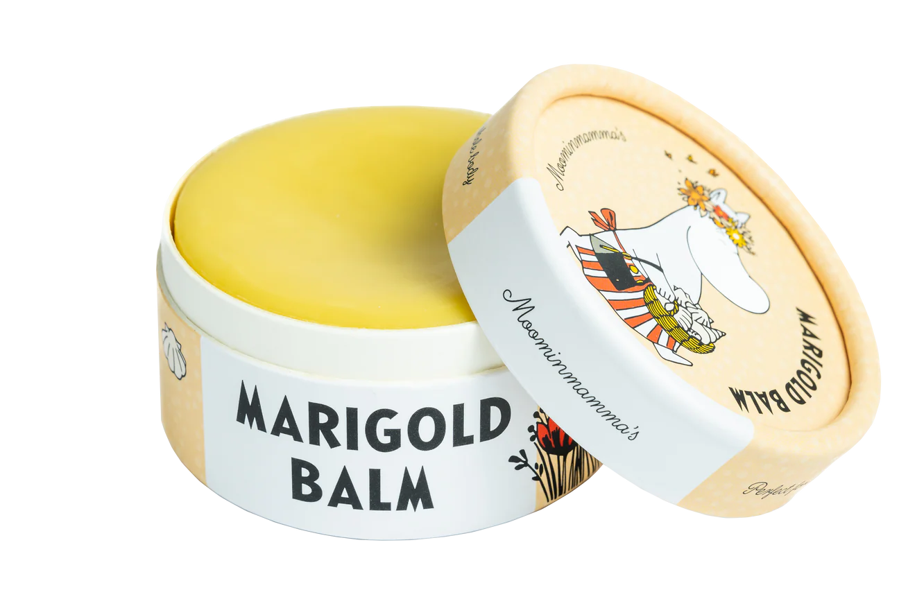 A round container of Gustaf & Linnea Moominmammas Marigold Balm with a partially open lid shows a Moomin character in a flower crown and striped scarf. The yellow natural skin balm comes in eco-friendly Gustaf & Linnea packaging.