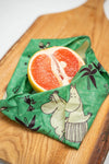 A halved grapefruit wrapped in a Gustaf & Linnea Moomin Beeswax Food Wrap rests on a wooden cutting board.