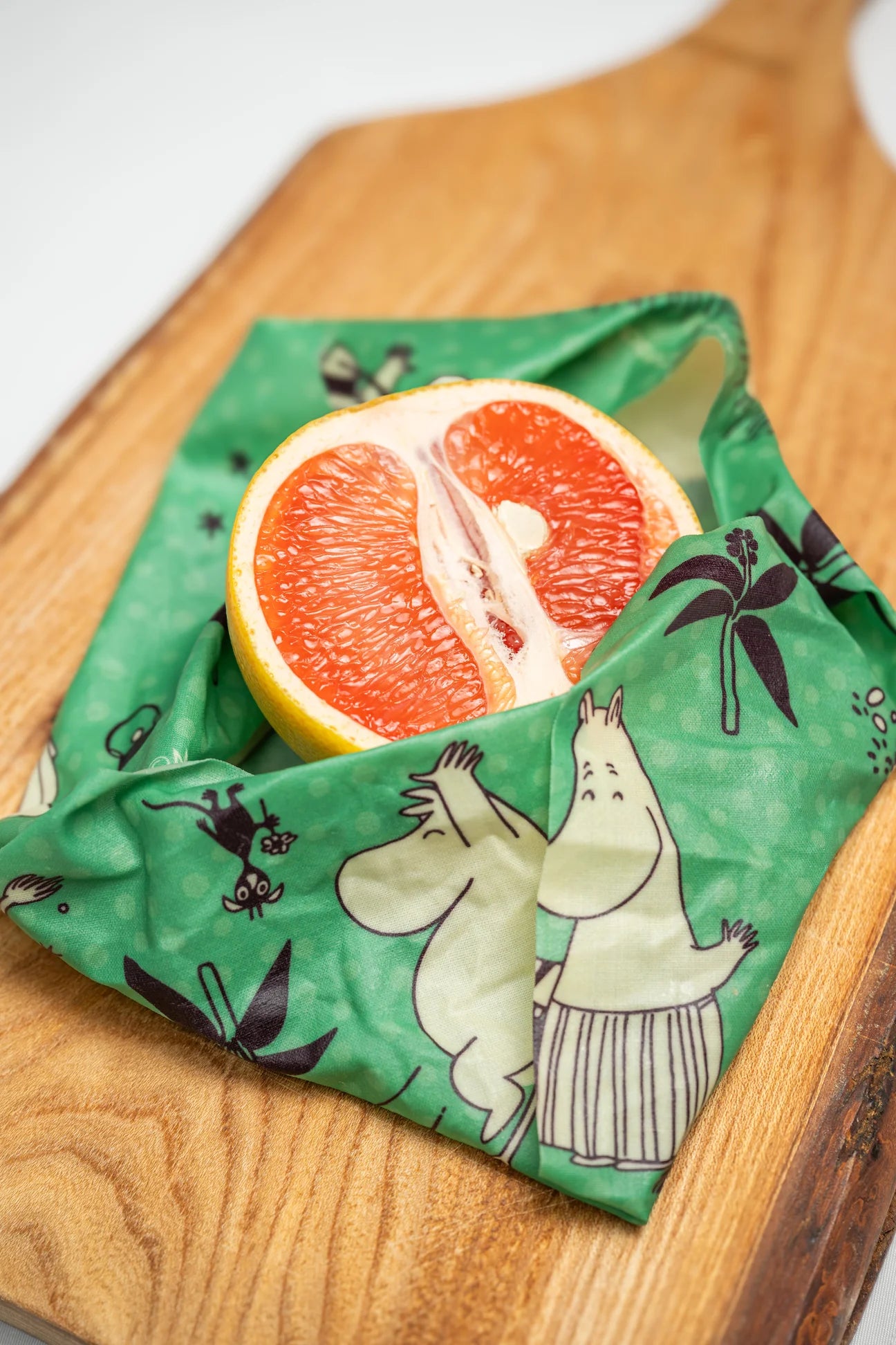 A halved grapefruit wrapped in a Gustaf & Linnea Moomin Beeswax Food Wrap rests on a wooden cutting board.