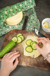 Two hands place cucumber slices on cheese-topped bread on a wooden board. A cucumber, cheese wheel, peeler, and bowl of cheese slices sit nearby, with a Gustaf & Linnea Moomin Beeswax Food Wrap adding a green touch in the background.