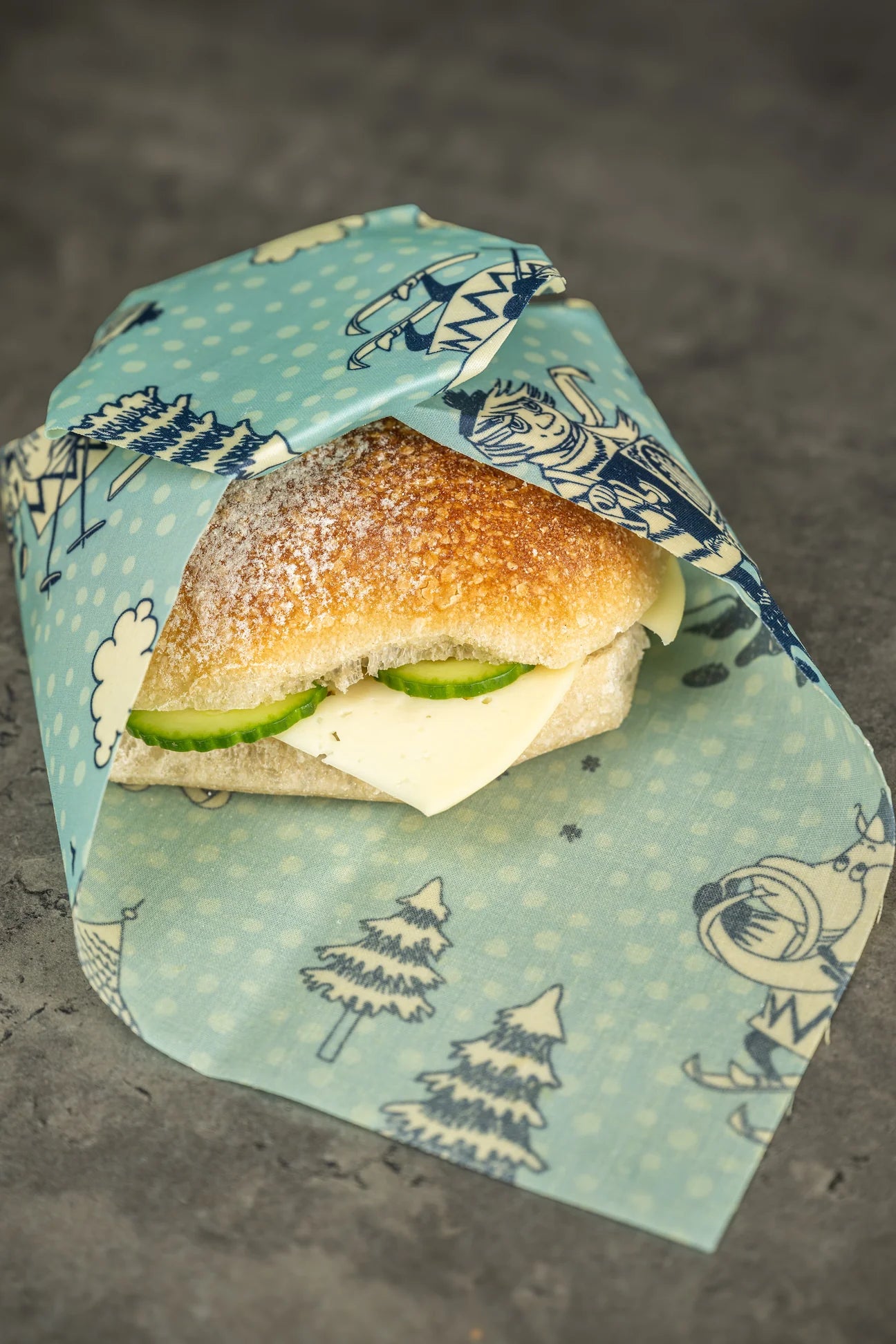 A cheese and cucumber sandwich in a crusty roll, partially wrapped in a Gustaf & Linnea Moomin Beeswax Food Wrap with winter patterns, sits on a gray surface.
