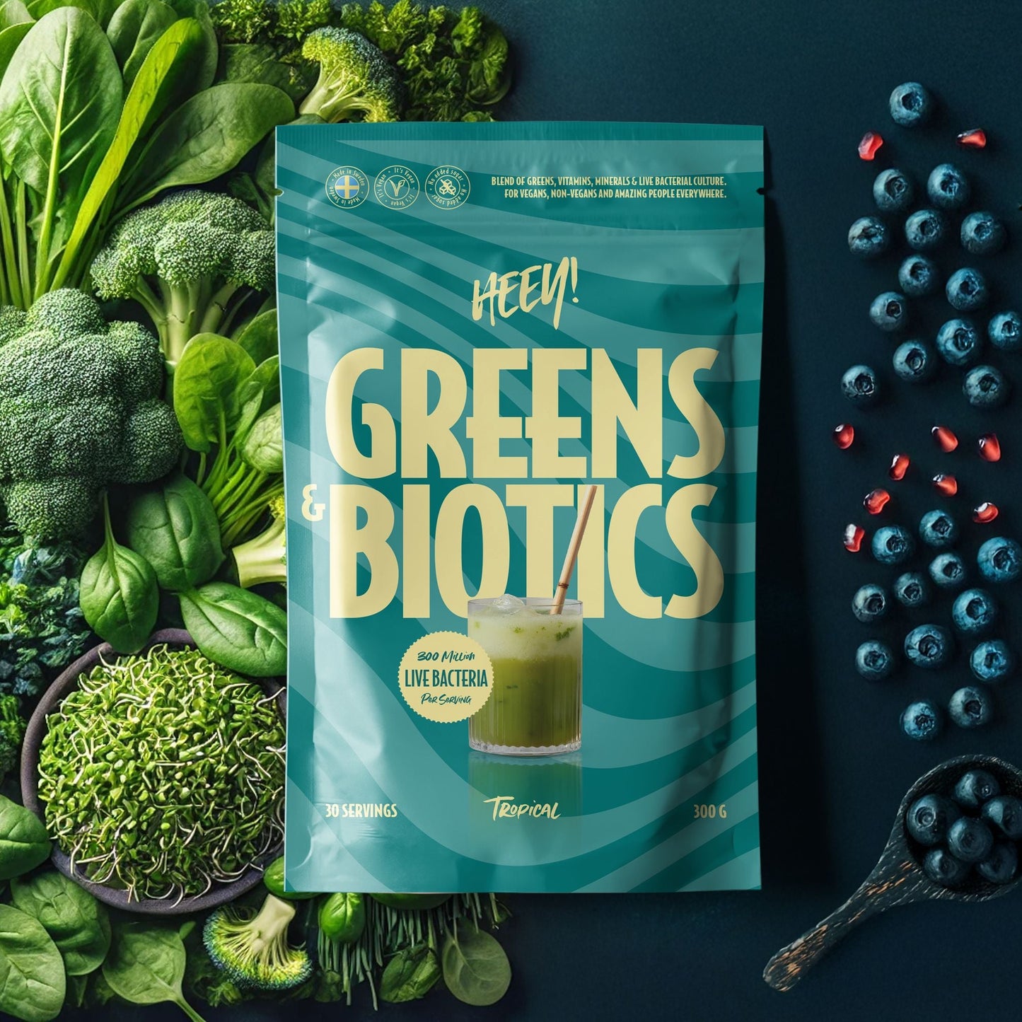 Package of 'HEEY! Greens & Biotics' surrounded by green vegetables and berries on a dark background
