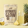 A stand-up pouch of Heey! Hydrate & Glow electrolyte powder in Pineapple Coconut flavor, styled with a glass of pale yellow iced drink, pineapple slices, coconut halves, and a whole pineapple on a warm yellow background.