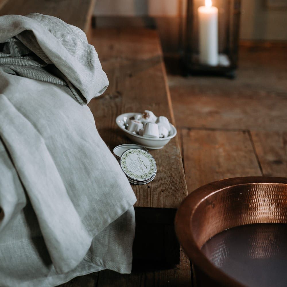 A cozy scene with a linen towel on a wooden bench, Kaurilan Sauna Lemongrass Footbath in a dish, a lit candle in a lantern, and a copper basin on a rustic wooden floor.