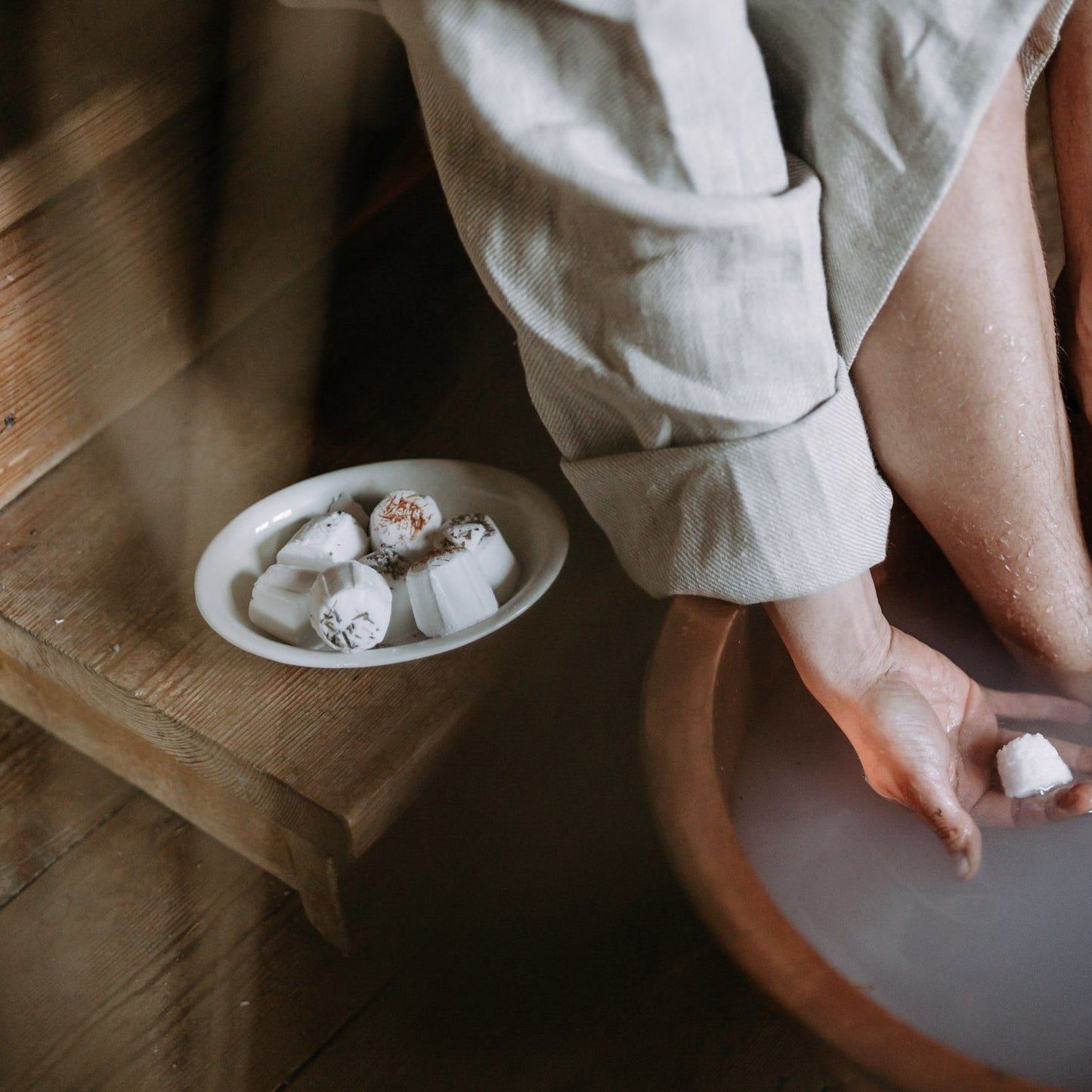 A person in a robe soaks their feet in cloudy water using Kaurilan Sauna Eucalyptus Footbath by Kaurilan Sauna, while a plate of natural cosmetics bath bombs rests on the wooden stairs nearby.