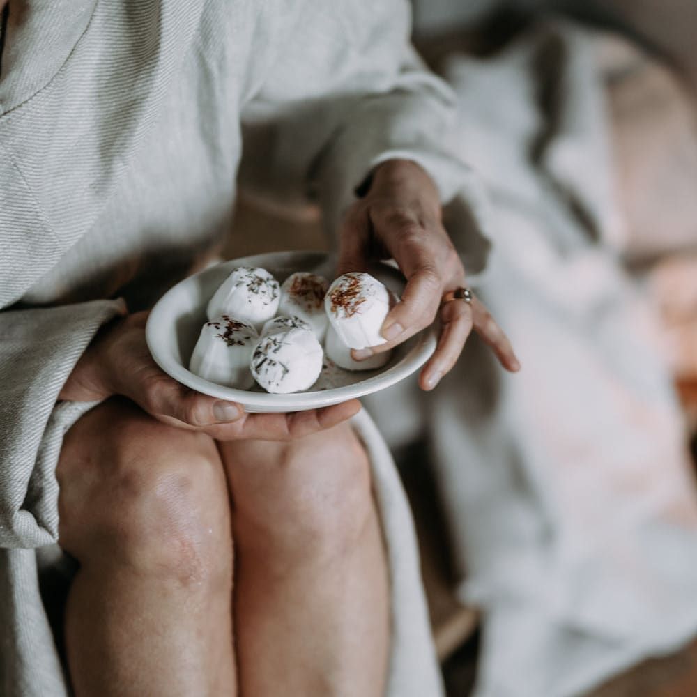 A person in a light robe sits with Kaurilan Sauna Eucalyptus Footbath by Kaurilan Sauna on their knees, holding one of the round, vegan, white desserts topped with small brown pieces.