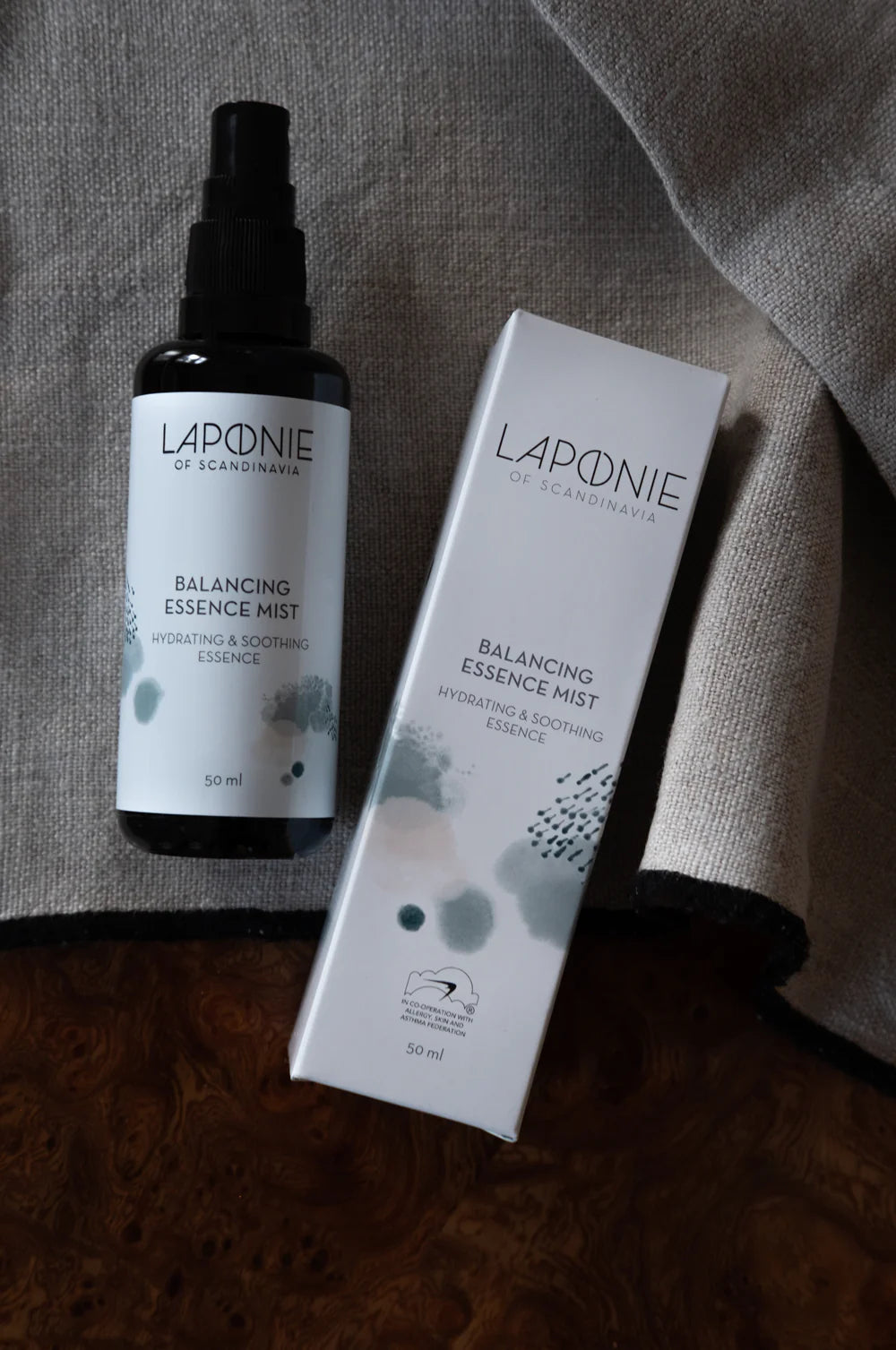 A bottle and box of Laponie of Scandinavia Balancing Essence Mist from Laponie of Scandinavia, a hydrating toner and face mist for sensitive skin, are shown on a grey fabric surface with a brown wooden edge.
