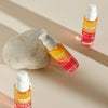 Three bottles of Lumene Nordic-C Arctic Berry Oil-Cocktail stand on a beige surface, one leaning against a smooth stone. The orange-to-pink ombre glass and white caps evoke skin glow from Arctic cloudberry extract.