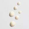 Six drops of Lumene Nordic-C Glow Awakening Eye Cream by Lumene, a creamy off-white vitamin C formula, are arranged diagonally on a white surface, with drop sizes decreasing from large to small.