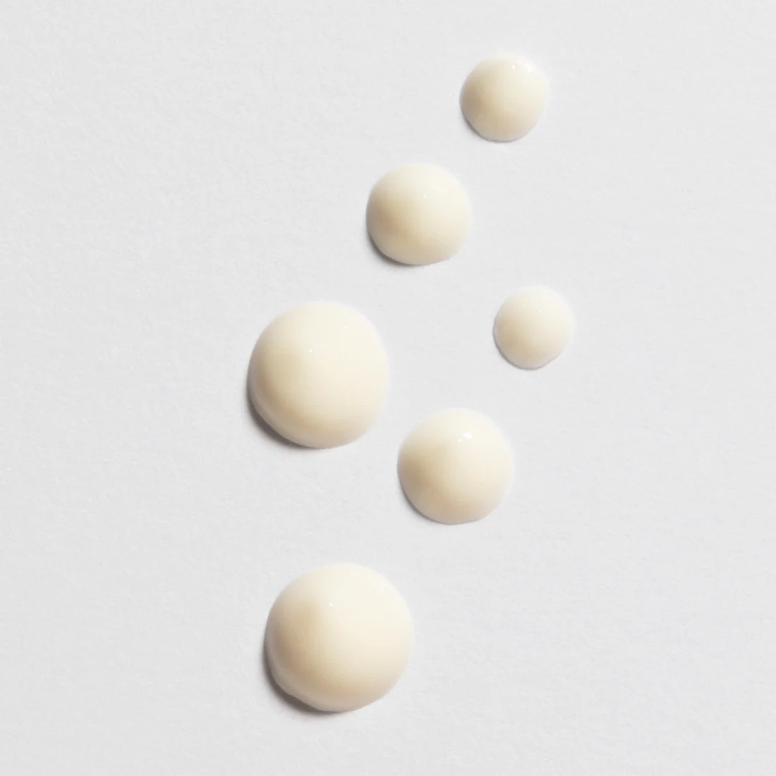 Six drops of Lumene Nordic-C Glow Awakening Eye Cream by Lumene, a creamy off-white vitamin C formula, are arranged diagonally on a white surface, with drop sizes decreasing from large to small.