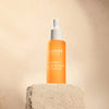 A bottle of Lumene Nordic-C Glow Boost Essence stands on a textured beige stone block against a soft beige background, featuring the brand Lumene and a white dropper cap.
