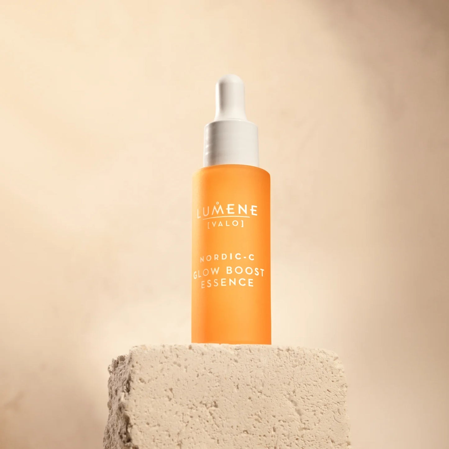 A bottle of Lumene Nordic-C Glow Boost Essence stands on a textured beige stone block against a soft beige background, featuring the brand Lumene and a white dropper cap.