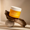 A yellow jar of Lumene Nordic-C Glow Moisturizer by Lumene, with a white lid, rests on driftwood atop smooth gray stone against a softly lit beige background.