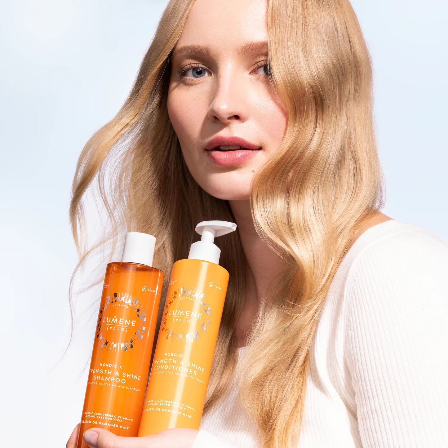 A woman with long, wavy blonde hair holds two orange bottles of Lumene Nordic-C Strength & Shine Shampoo by Lumene, standing against a light blue and white background. She wears a white top and looks toward the camera.
