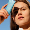 A woman holds a blue tube of Lumene Nordic Hydra Fresh Hydration Moisturizer SPF50 near her face, showcasing her daily hydration routine as sunlight casts a shadow across her forehead and eye.