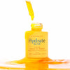 A dropper releases yellow serum into an open yellow bottle labeled Luonkos Bliss Facial Oil Serum by Luonkos, surrounded by a shiny pool of serum on a white background.