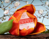 A red and orange tulip partially encircles a round tin of Luonkos Loved Special Edition Candy Red Lip Balm. White baby's breath flowers and a wooden surface appear in the background.